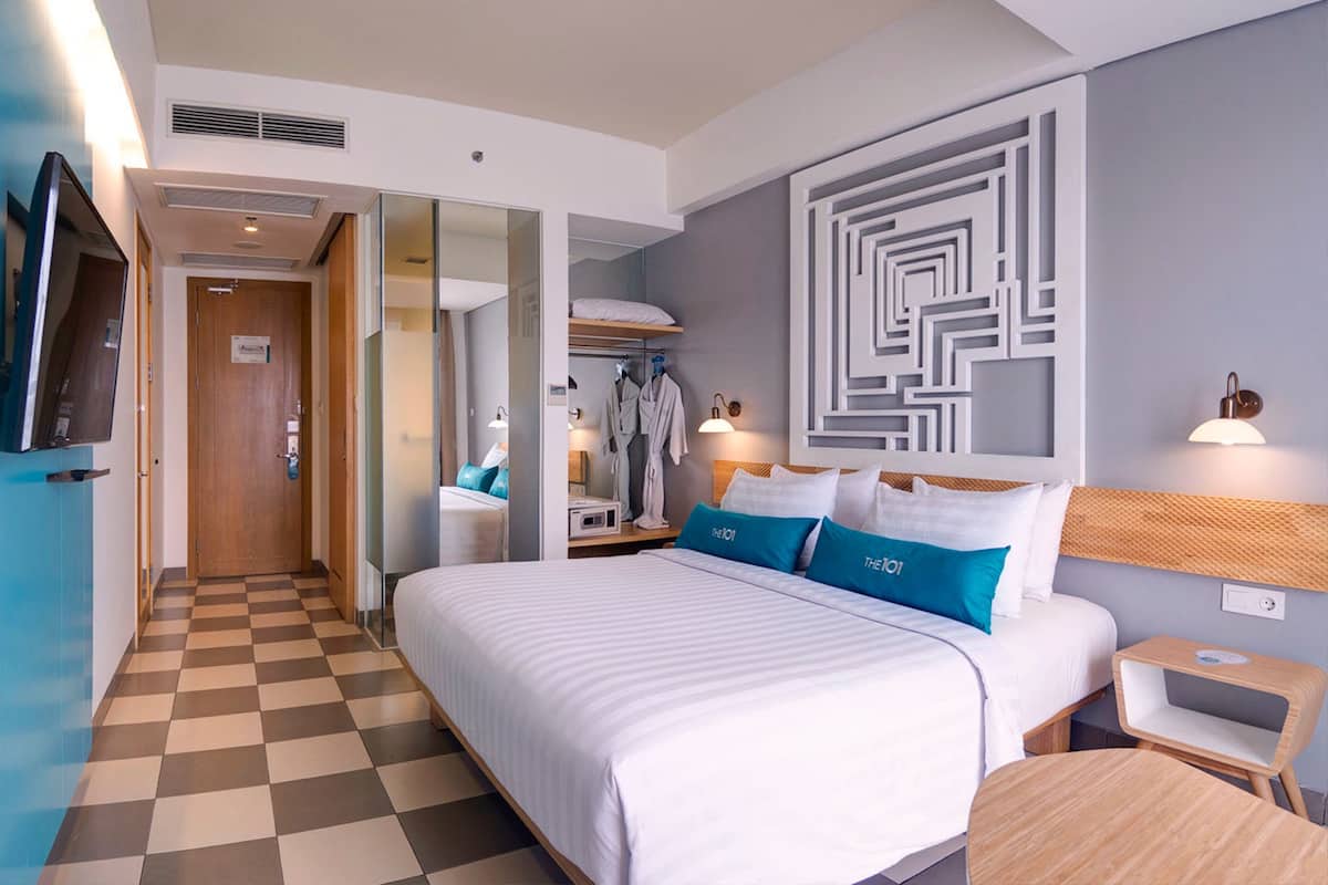 A hotel room with a checkered floor, a large bed with white and blue bedding, a wall-mounted TV, and a mirror.
