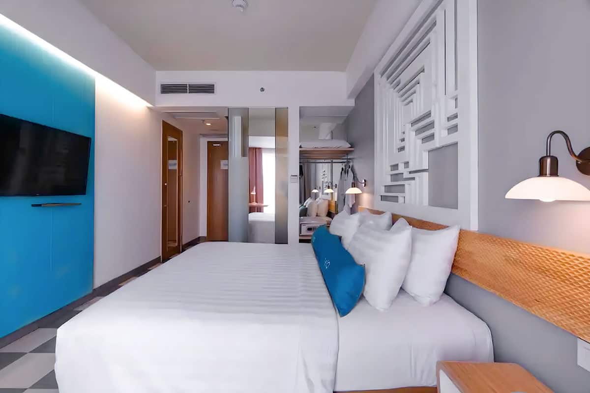 A modern hotel room with a large bed, a flat-screen TV mounted on the wall, a bedside table, and a lamp.