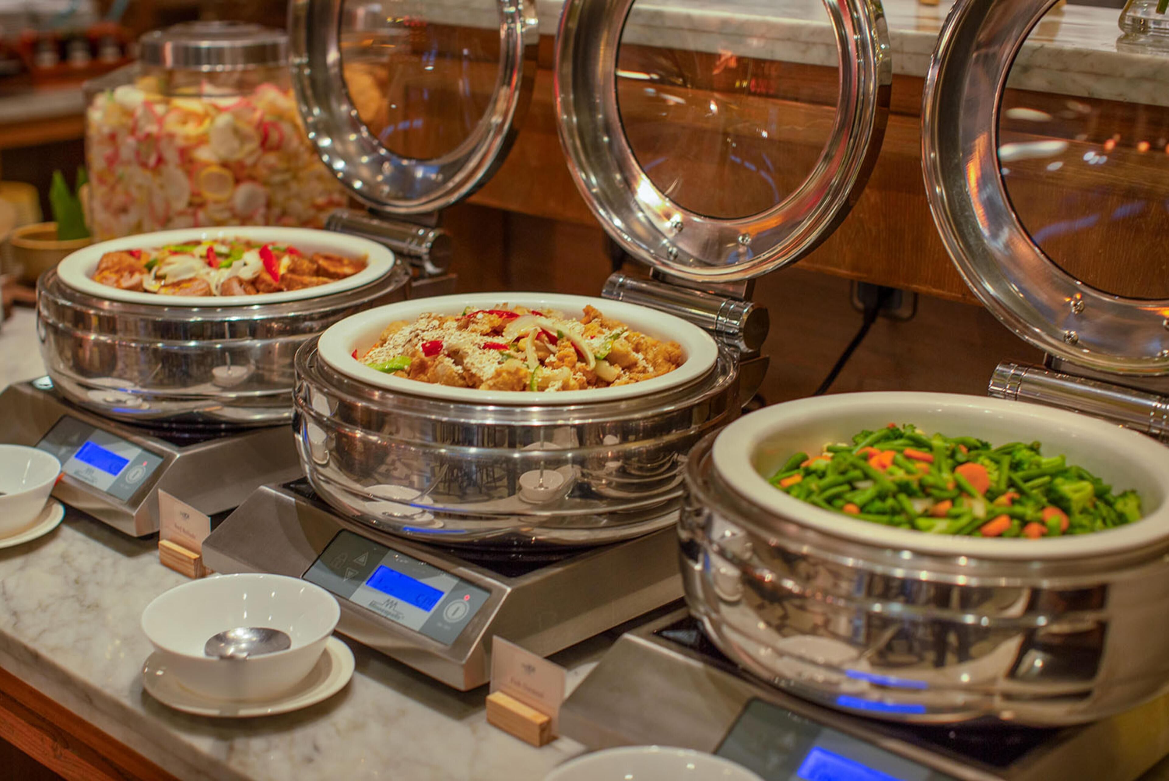 A buffet with various dishes in stainless steel bowls, some with lids, and digital scales for portion control.