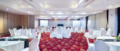 Un salón de banquetes con mesas redondas y sillas blancas, una alfombra roja y un escenario al frente.