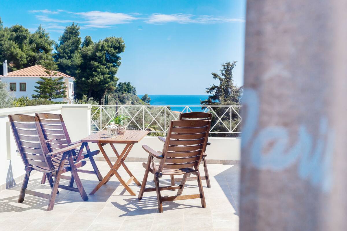 A terrace with two wooden chairs and a table, overlooking a coastal view with trees and a building in the background.