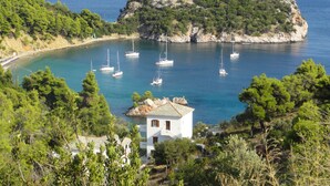 A coastal view with a small white building surrounded by lush greenery, a rocky island in the background, and several sailboats anchored in the water. - Stafylos Suites Boutique Hotel (Skopelos)