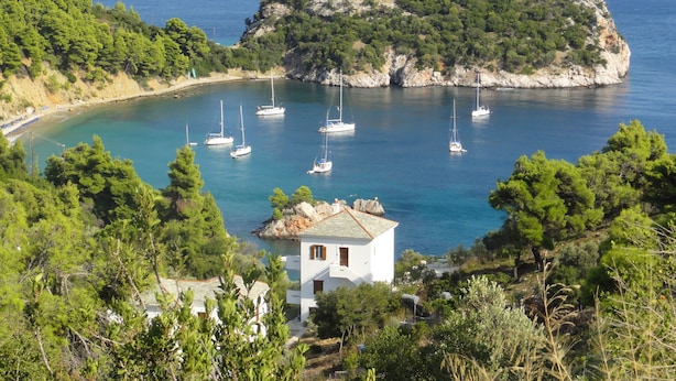 Une vue sur la côte, avec un petit bâtiment blanc entouré d’une végétation luxuriante, une île rocheuse en arrière-plan et plusieurs voiliers ancrés dans l’eau.