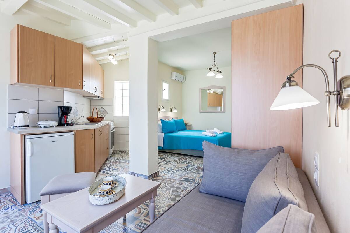 A compact living space with a kitchenette, a sofa, a small table, and a bed with blue bedding.