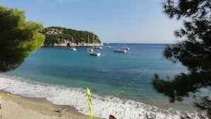 Une plage bordée d’une eau turquoise et limpide, des bateaux ancrés au large et un promontoire rocheux couvert de végétation.