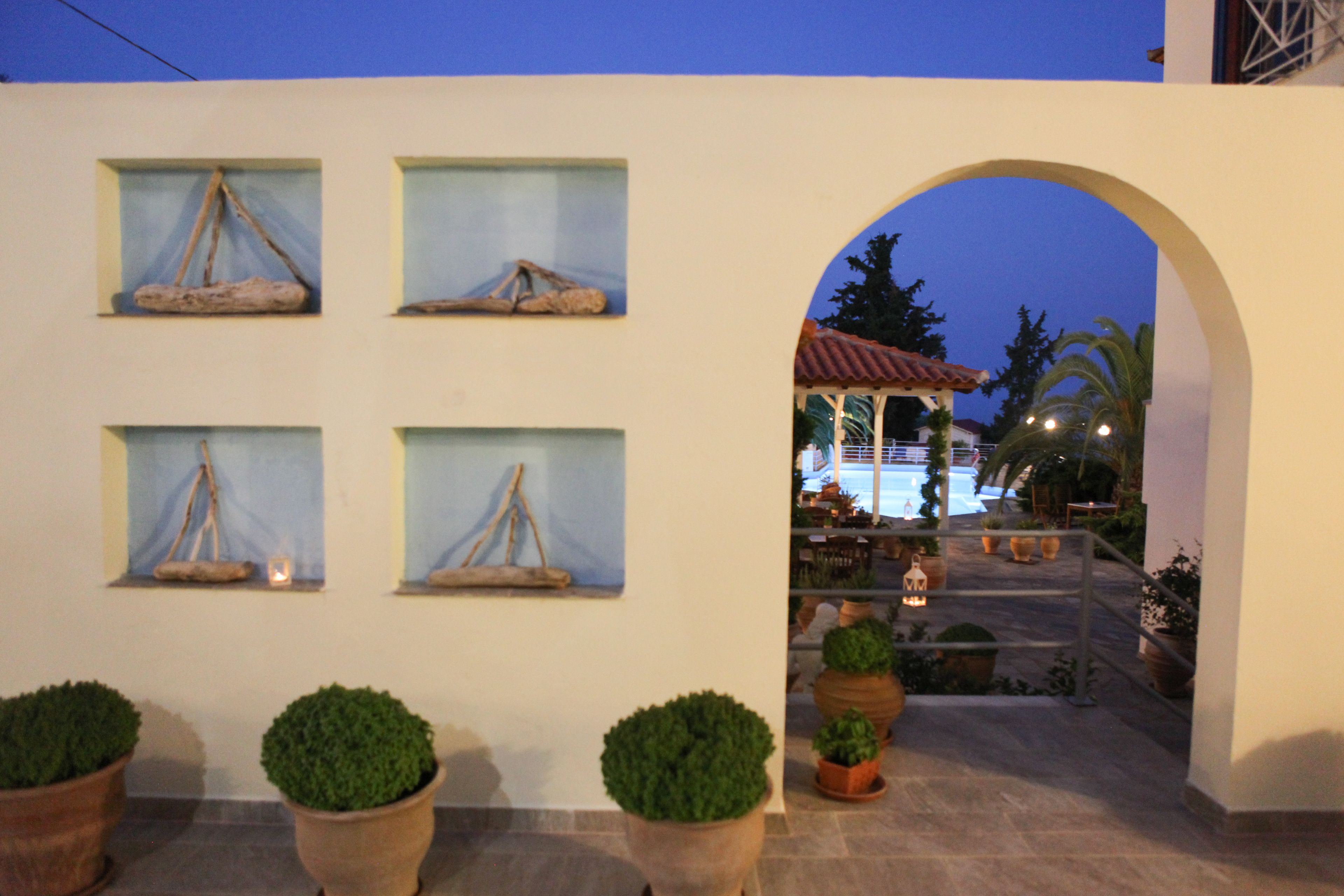 A building facade with multiple windows, some with decorative elements, and a central archway leading to an outdoor area with potted plants and a view of a building with a red roof.