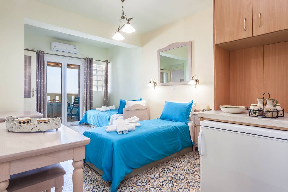 A hotel room with two beds, a dining table, a kitchen area, and a sliding glass door leading to a balcony.