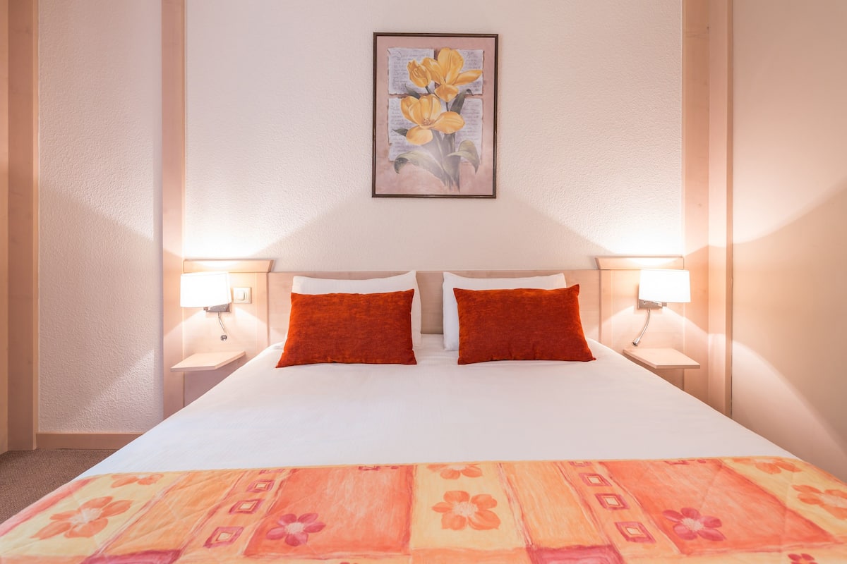 Une chambre d’hôtel avec un grand lit, deux oreillers rouges, une couvre-lit à motifs floraux et un tableau encadré accroché au mur.