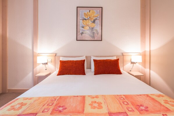 Una habitación de hotel con una cama grande, dos almohadas rojas, una colcha estampada con motivos florales y un cuadro en la pared.