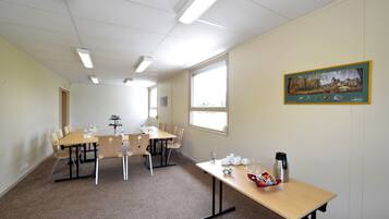 Un salón de conferencias con una larga mesa, sillas y un proyector. Hay una ventana con persianas, un cuadro en la pared y una mesa con vasos y un termo.