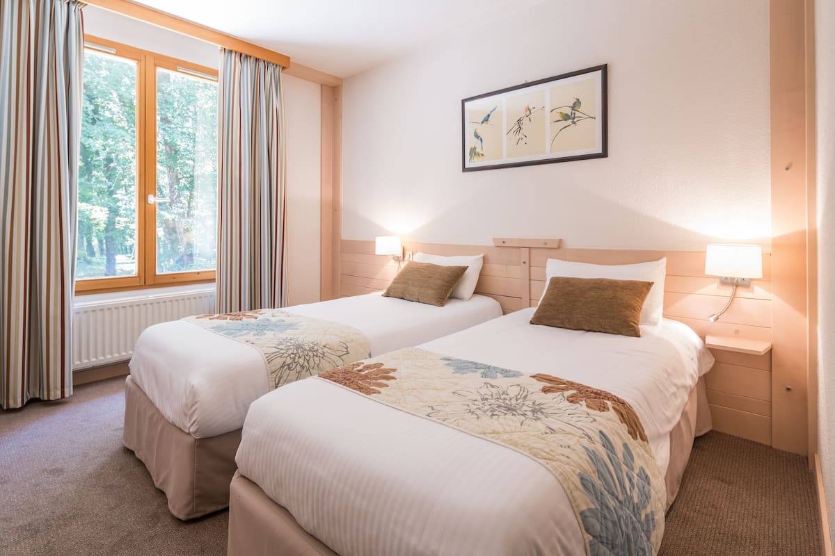 Une chambre d’hôtel avec deux lits, une fenêtre avec des rideaux, un tableau accroché au mur et des lampes de chevet.