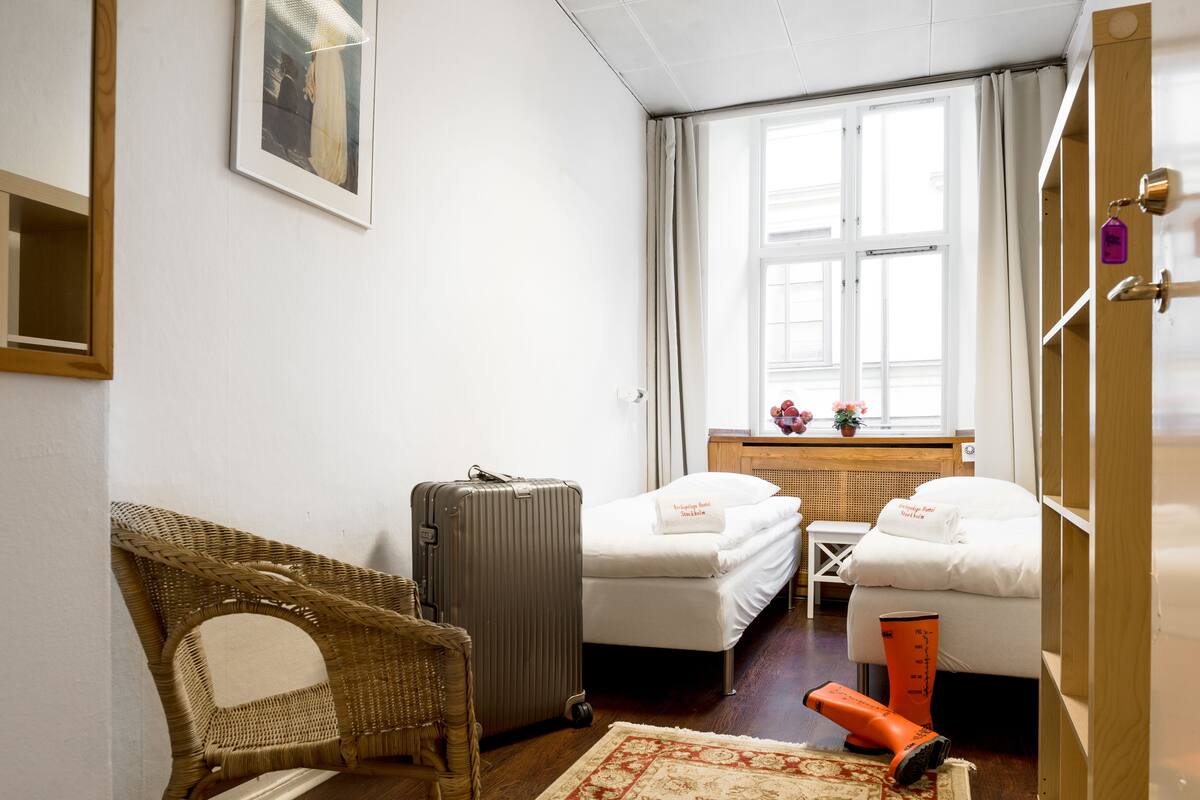 A hotel room with two single beds, a wicker chair, a suitcase, a small table, and a window with curtains.