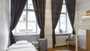 A hotel room with a bed, a suitcase, a small table with a remote, a wicker chair with cushions, a bunk bed, and a window with curtains. - Archipelago Hostel Old Town (Stockholm)