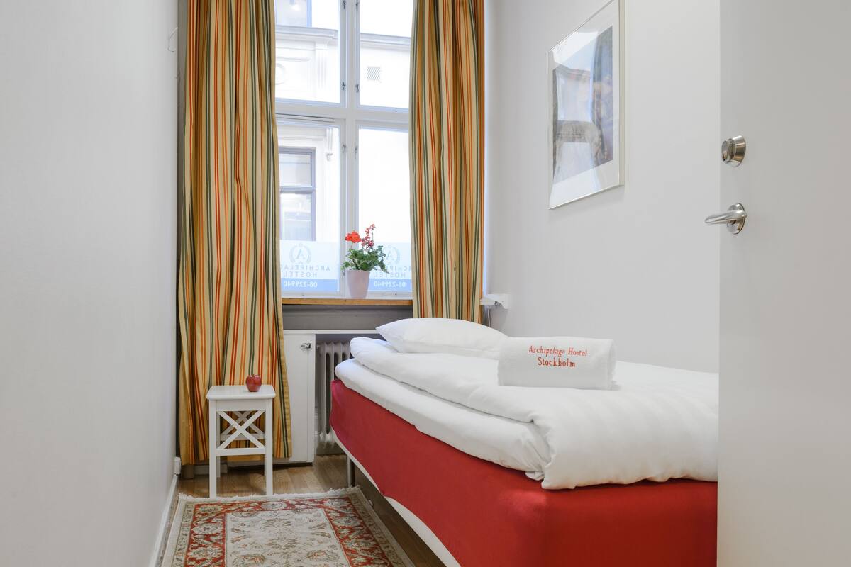 A hotel room with a bed, a window with curtains, a radiator, a small table with a plant, and a framed picture on the wall.