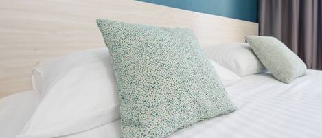 Una cama bien tendida con sábanas blancas y dos almohadas, una con un estampado azul verdoso y la otra blanca lisa, frente a una pared azul y un cabecero de madera.