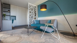 A modern living room with a teal sofa, a black coffee table, a white chair, a floor lamp, and a decorative wall panel. - Hotel Kolovare (Zadar)