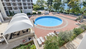 A pool area with lounge chairs and a covered seating area, surrounded by greenery and a building with a modern design. - Hotel Kolovare (Zadar)