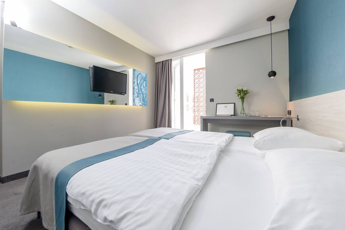 A hotel room with a large bed, a flat-screen TV mounted on the wall, a small table with a vase of flowers, and a window with curtains.
