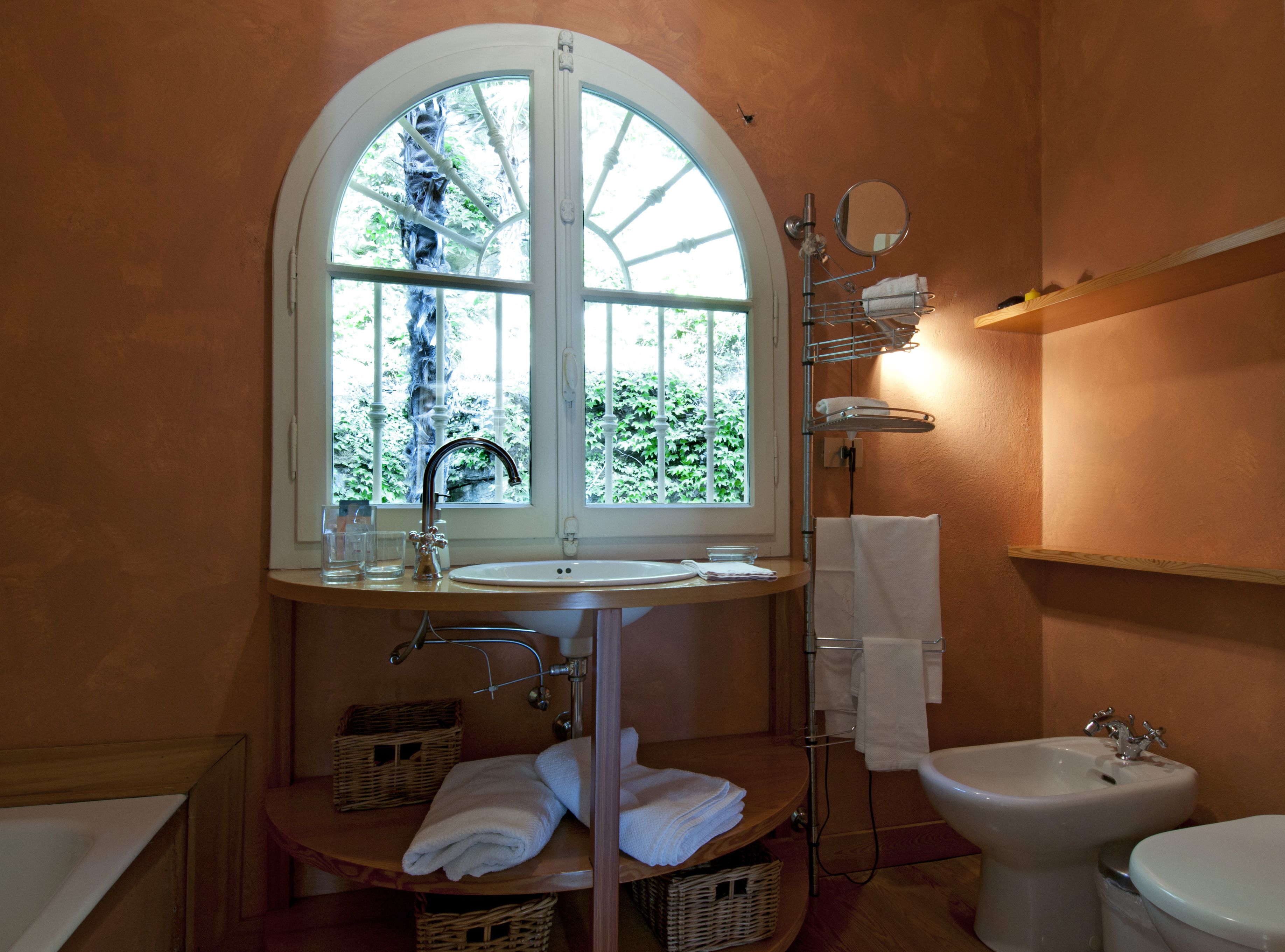 A bathroom with a large arched window, a sink, a bidet, and a toilet. There are baskets and towels on the counter.