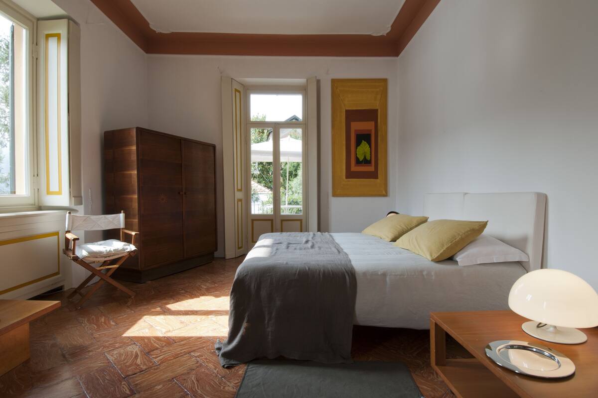A bedroom with a bed, a chair, a wardrobe, a bedside table with a lamp, and a window.