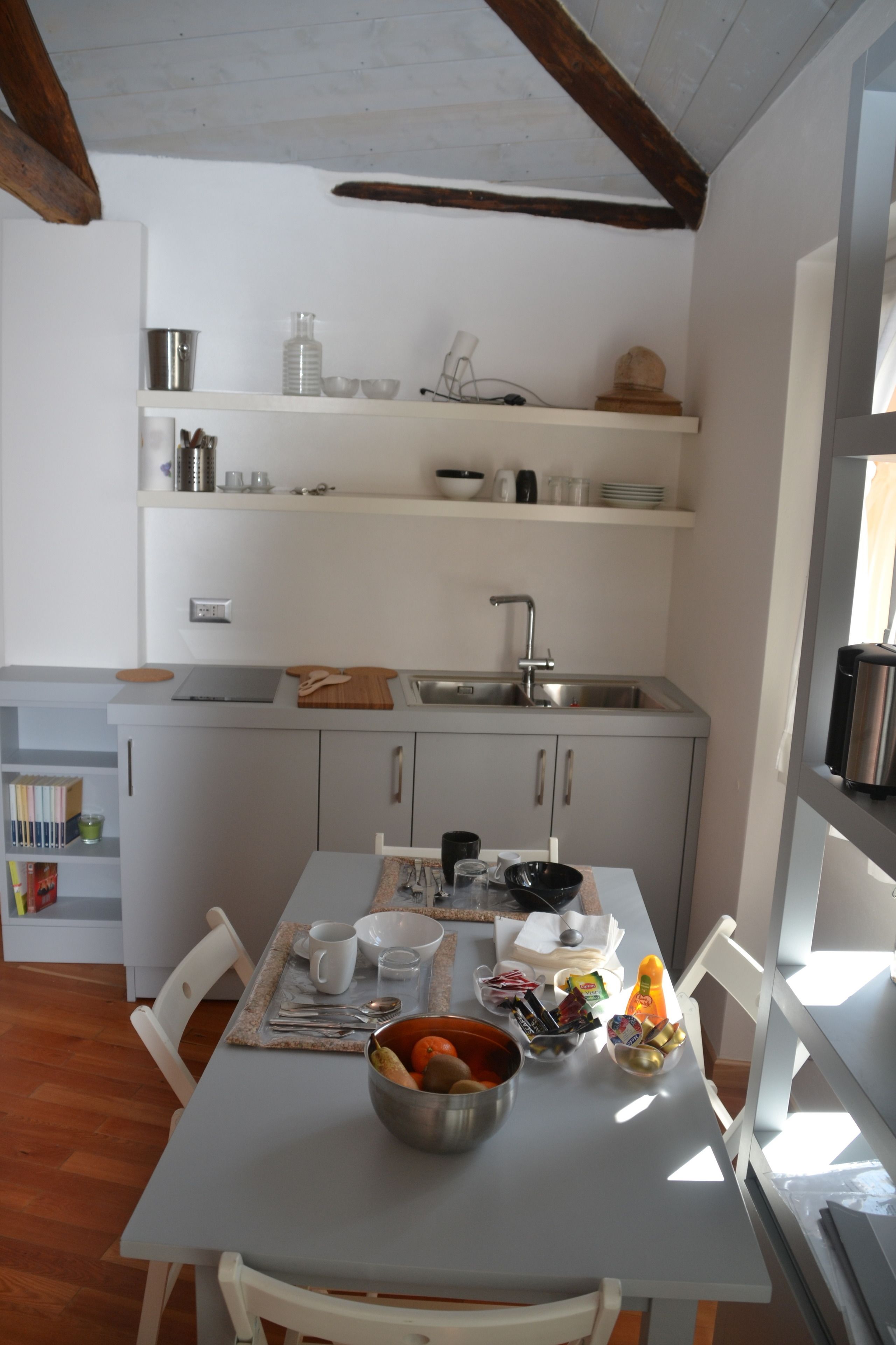 Une cuisine compacte avec un évier, des placards blancs et une table à manger avec des chaises. Des étagères au-dessus de l’évier servent à ranger divers ustensiles de cuisine.