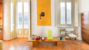 A room with a wooden floor, a bench with a coffee pot, cups, and a green box, a folding chair, and a wall-mounted artwork. - Villa Nina Relais Boutique B&B (Carate Urio)