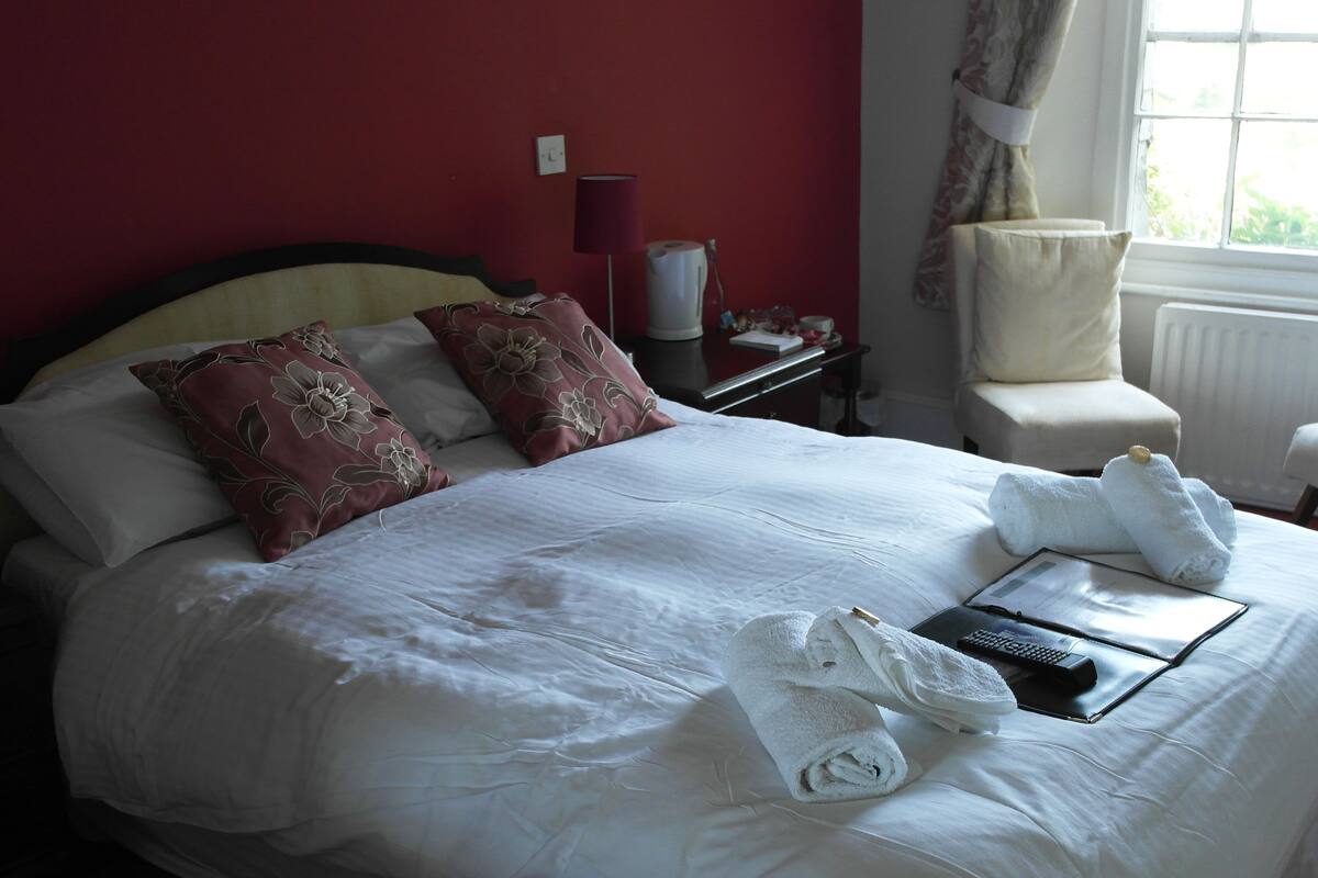 A neatly made bed with white linens and floral pillows, a bedside table with a lamp and a cup, and a window with curtains.