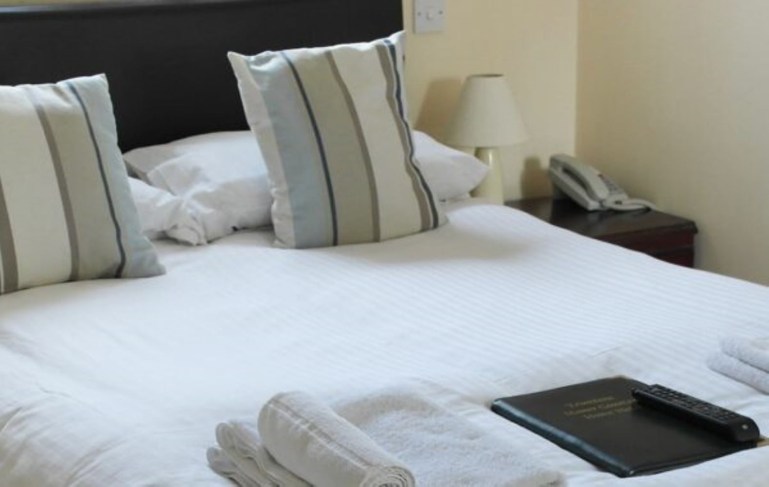 A neatly made bed with pillows, a folded towel, and a remote control on a book.