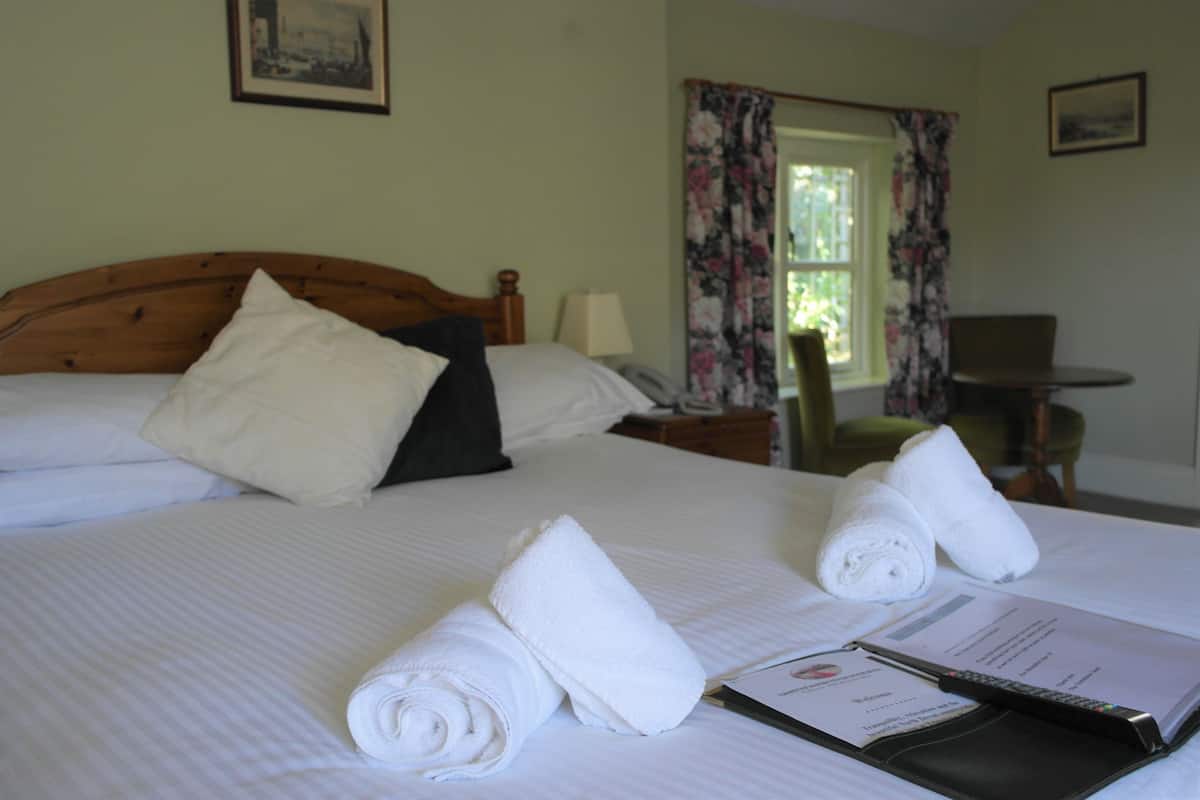A neatly made bed with rolled white towels, a bedside table with a lamp, and a chair in a room with floral curtains and a framed picture on the wall.