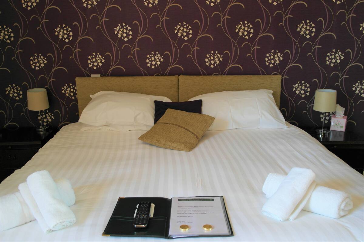A neatly made bed with white linens, a brown pillow, and a remote control on a black tray. There are rolled towels on the bed and a booklet with a menu on the tray.