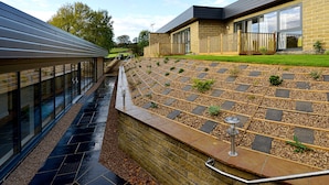 Een modern gebouw met een glazen gevel, een stenen keerwand en een aangelegde tuin met een houten terras.