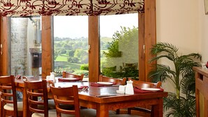 Een eetgedeelte met een houten tafel en stoelen, uitzicht op een groen landschap door grote ramen, en een potplant aan de rechterkant.
