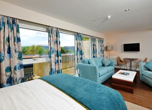 Une chambre d’hôtel moderne, dotée d’un grand lit, d’un canapé, d’une table basse et d’un téléviseur. La chambre dispose de grandes fenêtres avec des rideaux et offre une vue sur l’extérieur.