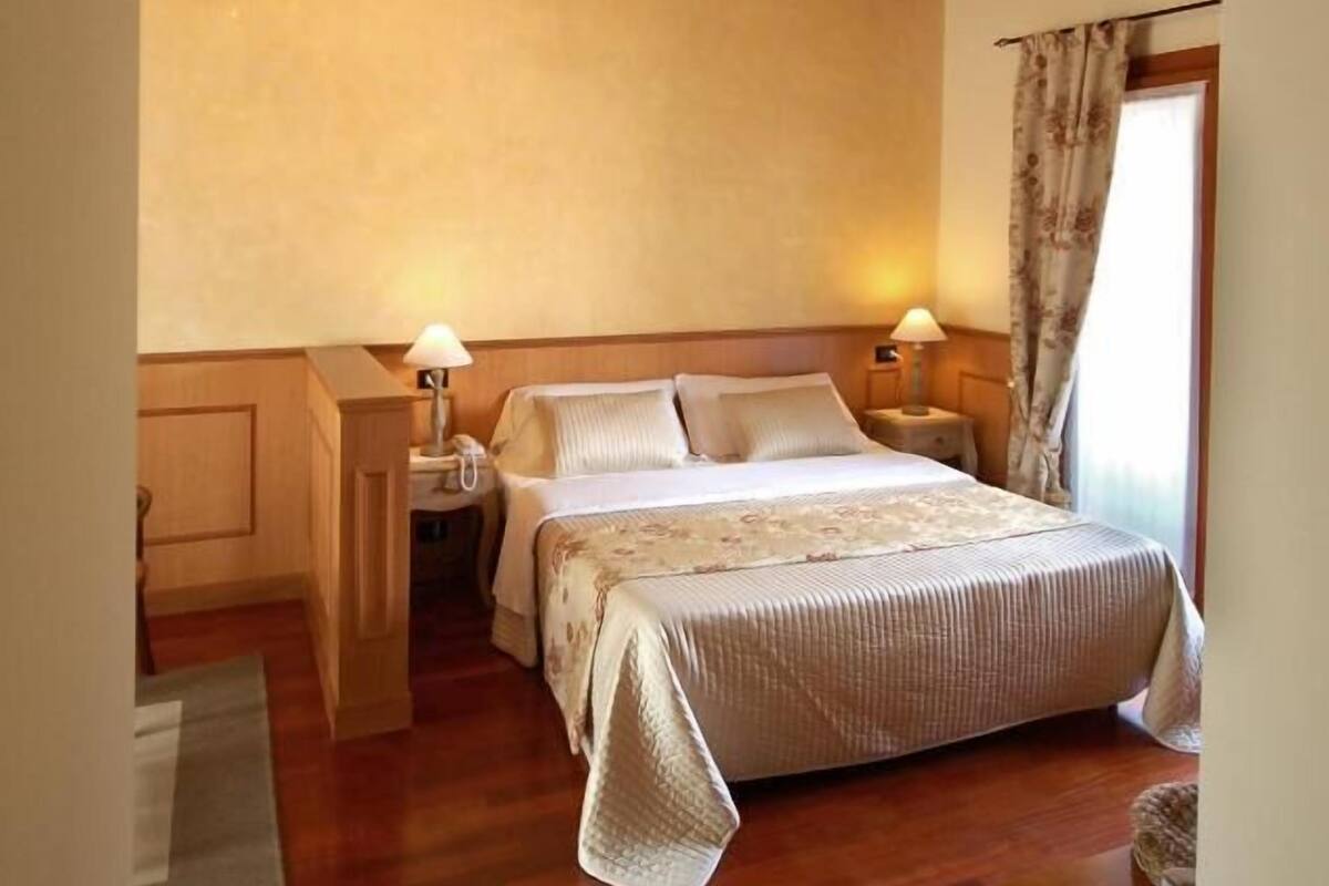 A bedroom with a wooden ceiling, a chandelier, a bed with a floral bedspread, bedside lamps, and a window with curtains.