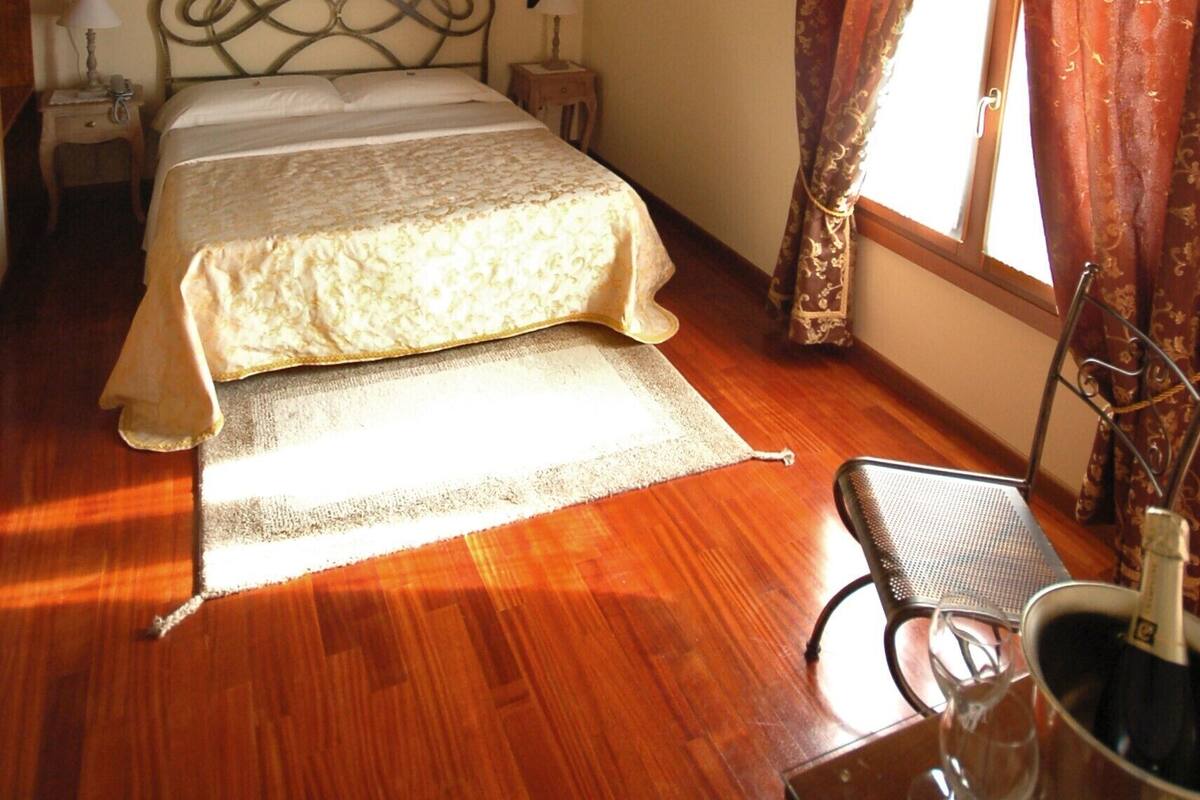 A bedroom with a wooden ceiling, a bed with a headboard, two bedside tables with lamps, a window with curtains, and a chair with a bottle of champagne.