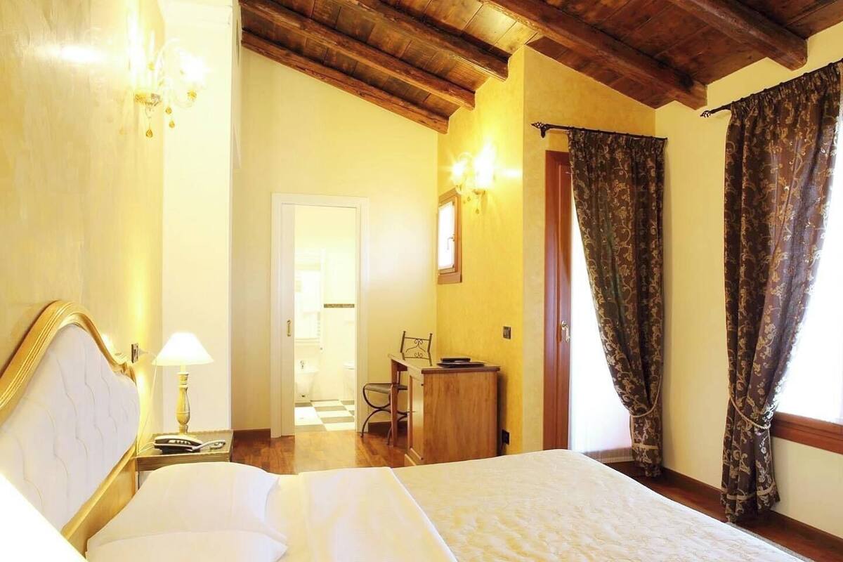 A hotel room with a large bed, wooden ceiling beams, a desk with a chair, and a view of the outdoors through curtains.