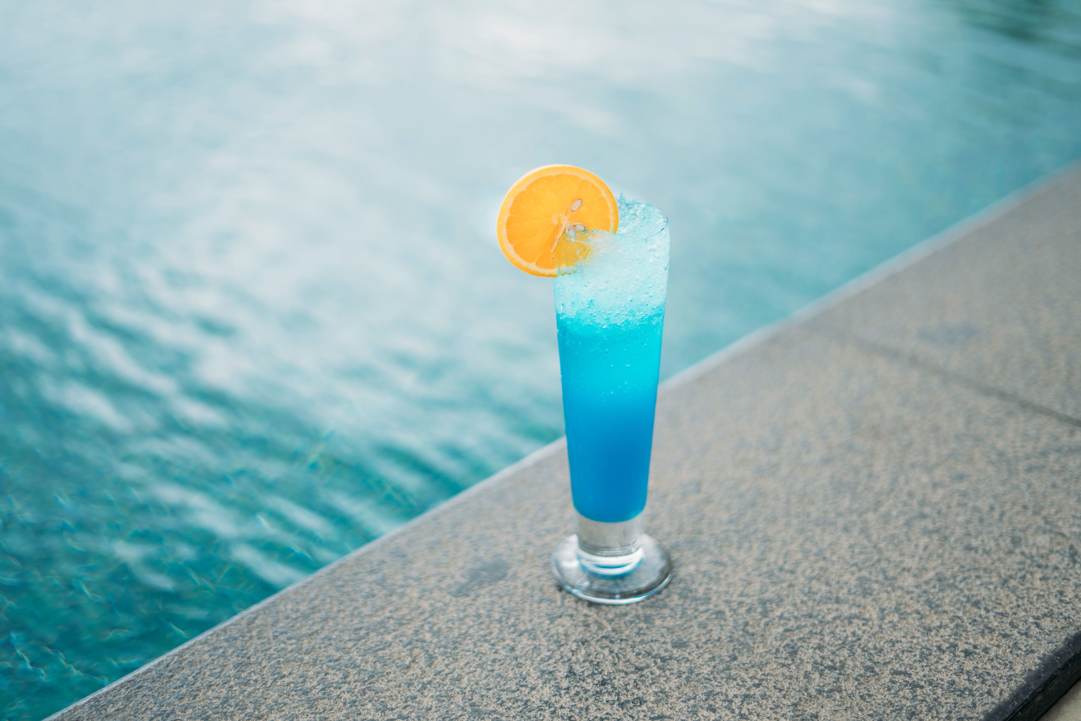 Un vaso alto con líquido azul y una rodaja de naranja, sobre un borde de piedra junto a una masa de agua.