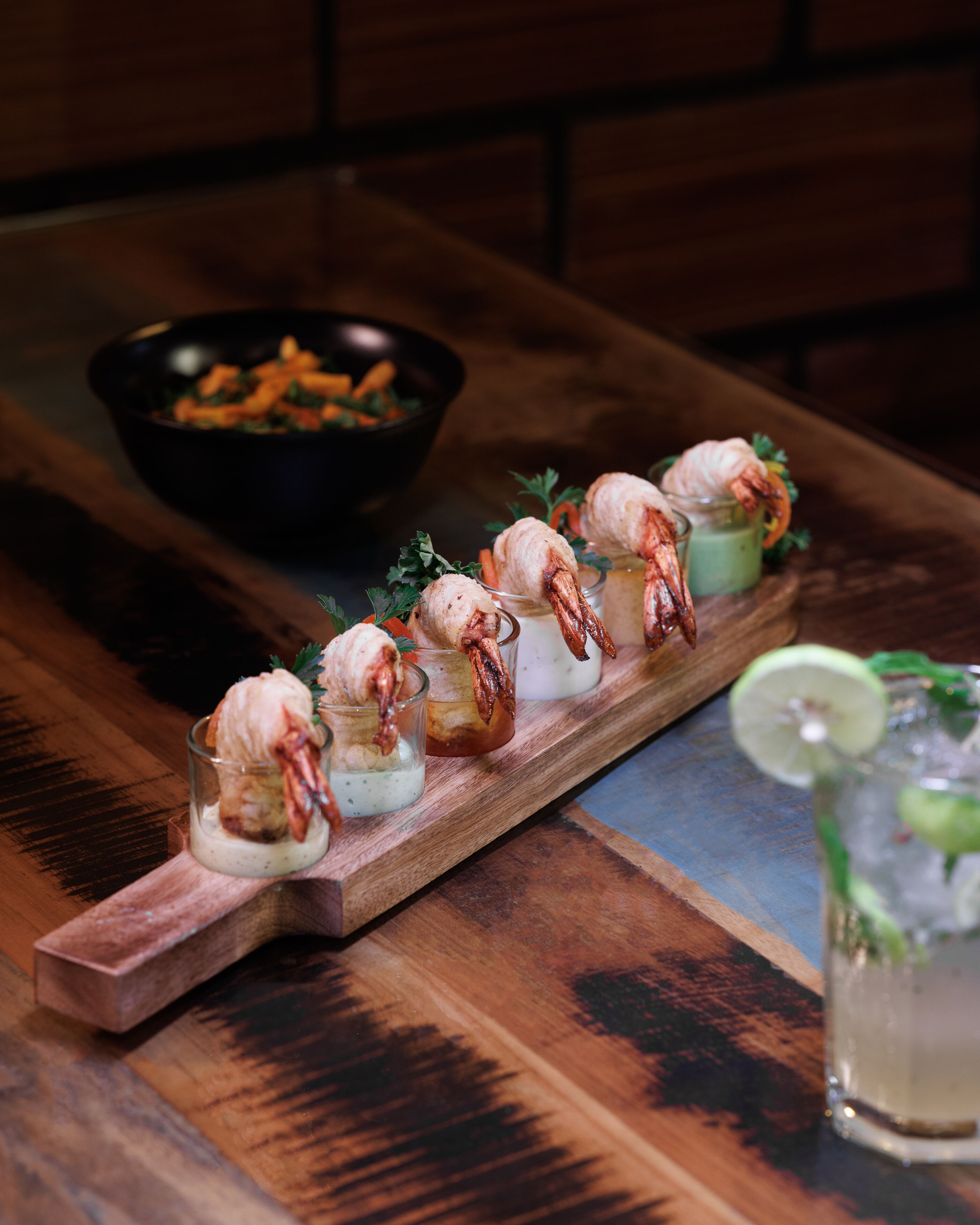 A wooden table with a glass tray holding shrimp appetizers and a bowl of vegetables in the background.