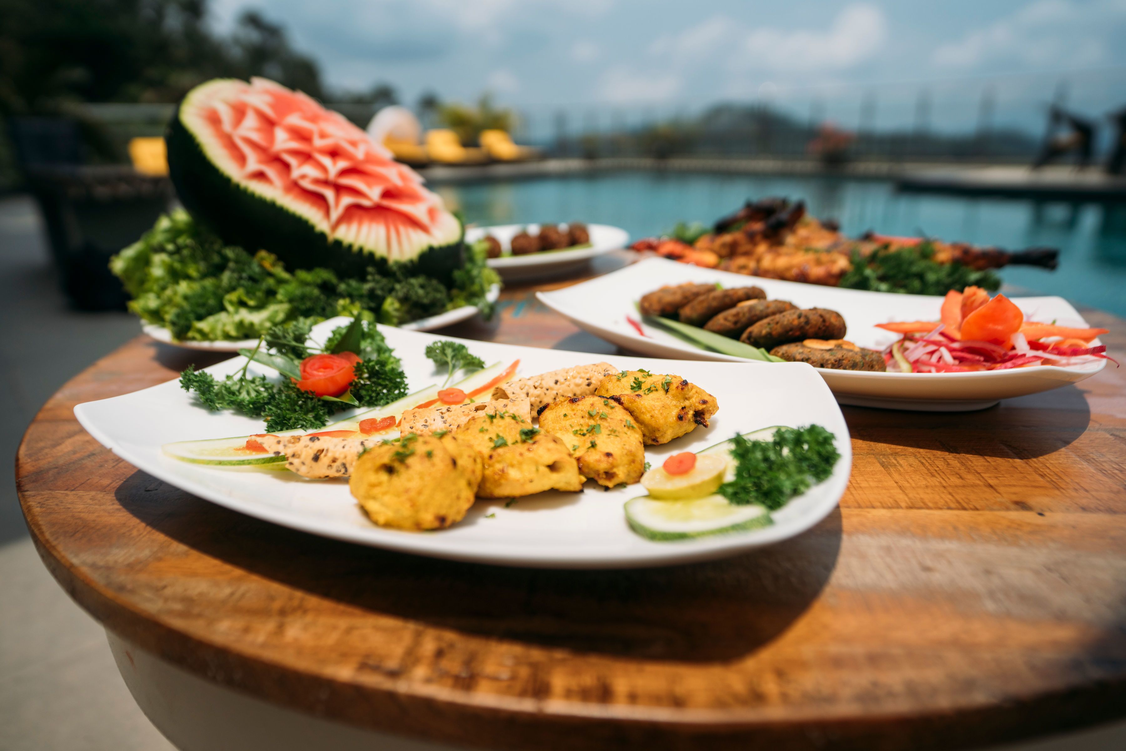 Diverse gerechten op een houten tafel bij een zwembad, waaronder een uit een watermeloen gesneden bloem en verschillende borden met eten.