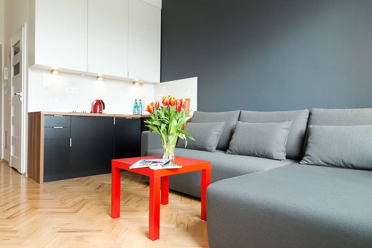 Una sala de estar moderna con un sofá gris, una mesa roja y una cocina al fondo.