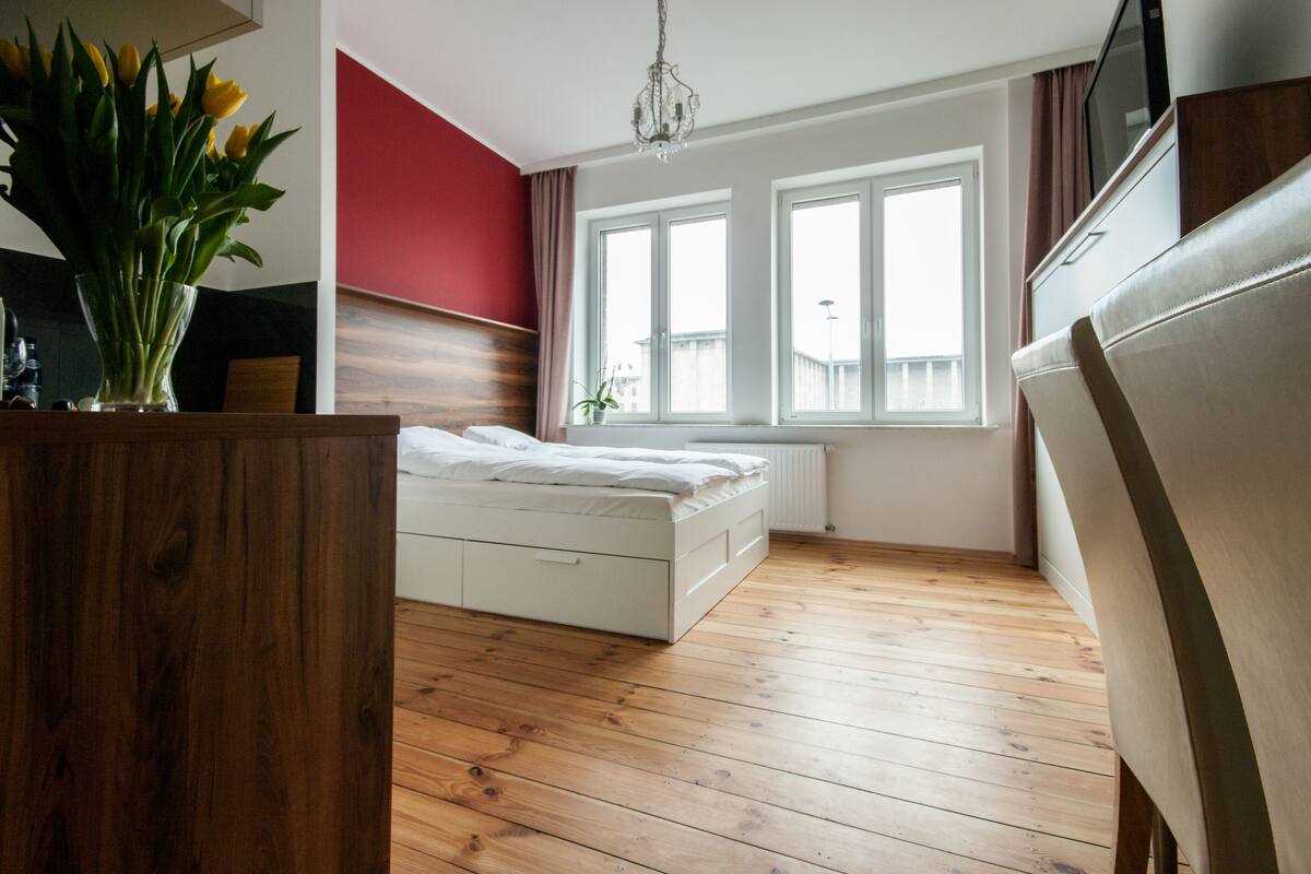 Un dormitorio moderno con suelo de madera, cama blanca, pared de acento roja y ventana con vistas.