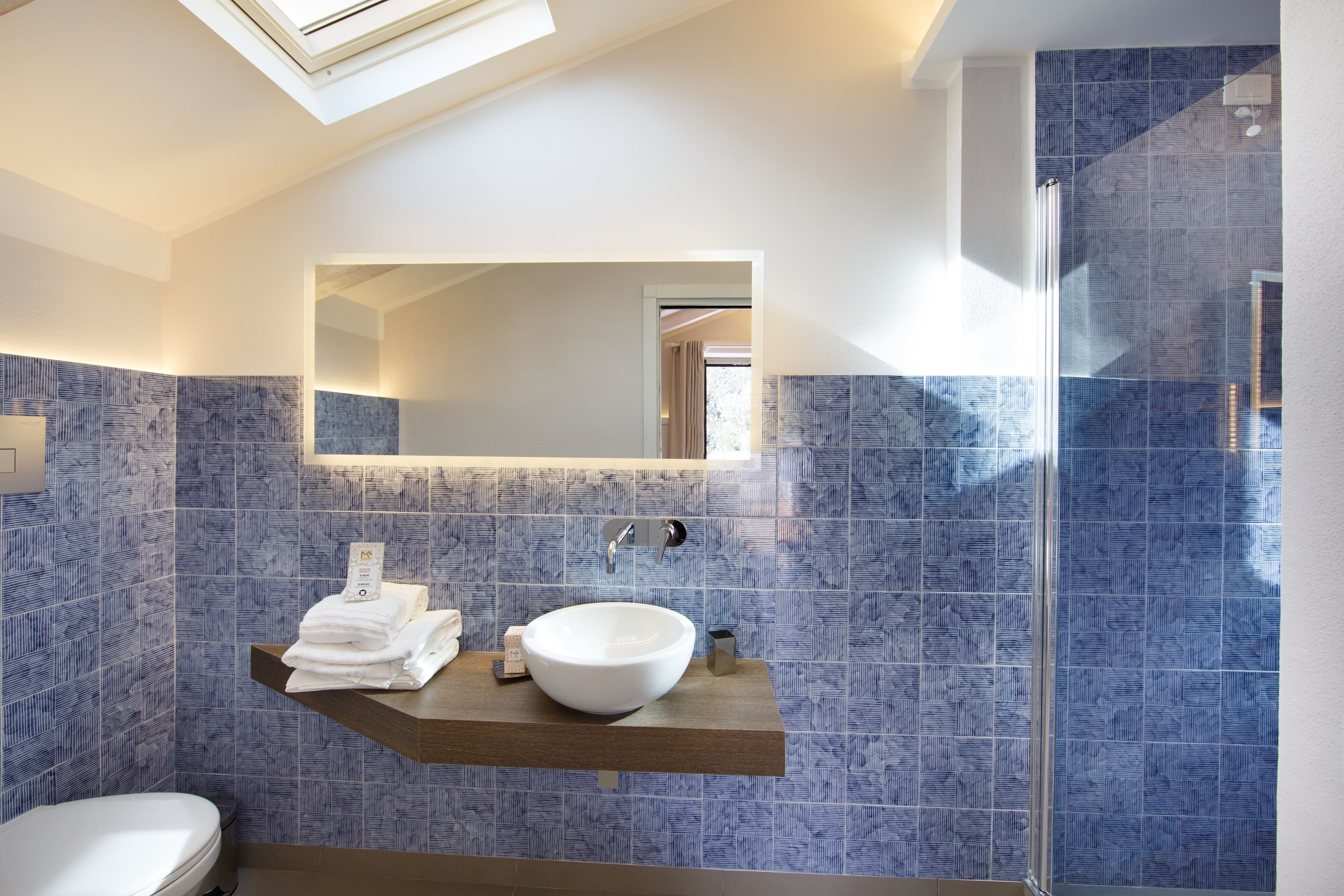 Een moderne badkamer met dakraam, grote spiegel, wit wastafelmeubel en blauwe betegelde vloer.