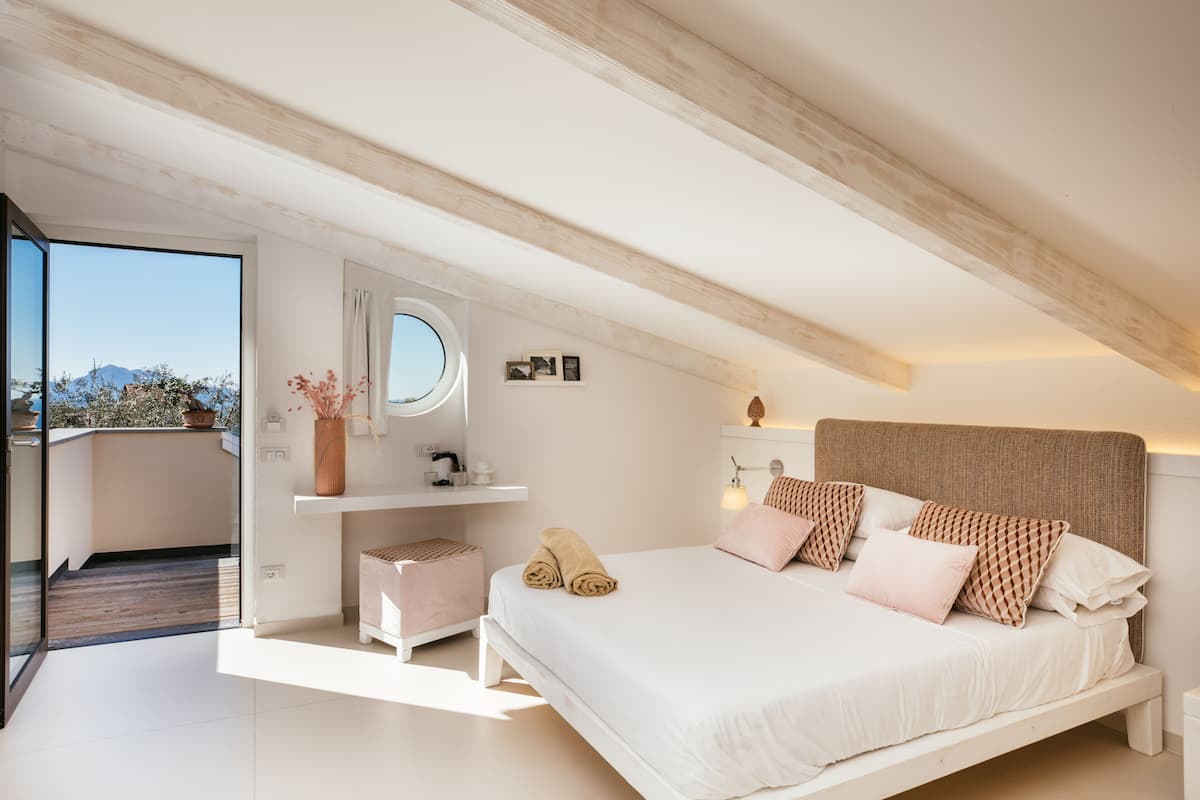 A bedroom with a large bed, a bedside table, a mirror, and a view of the outdoors.