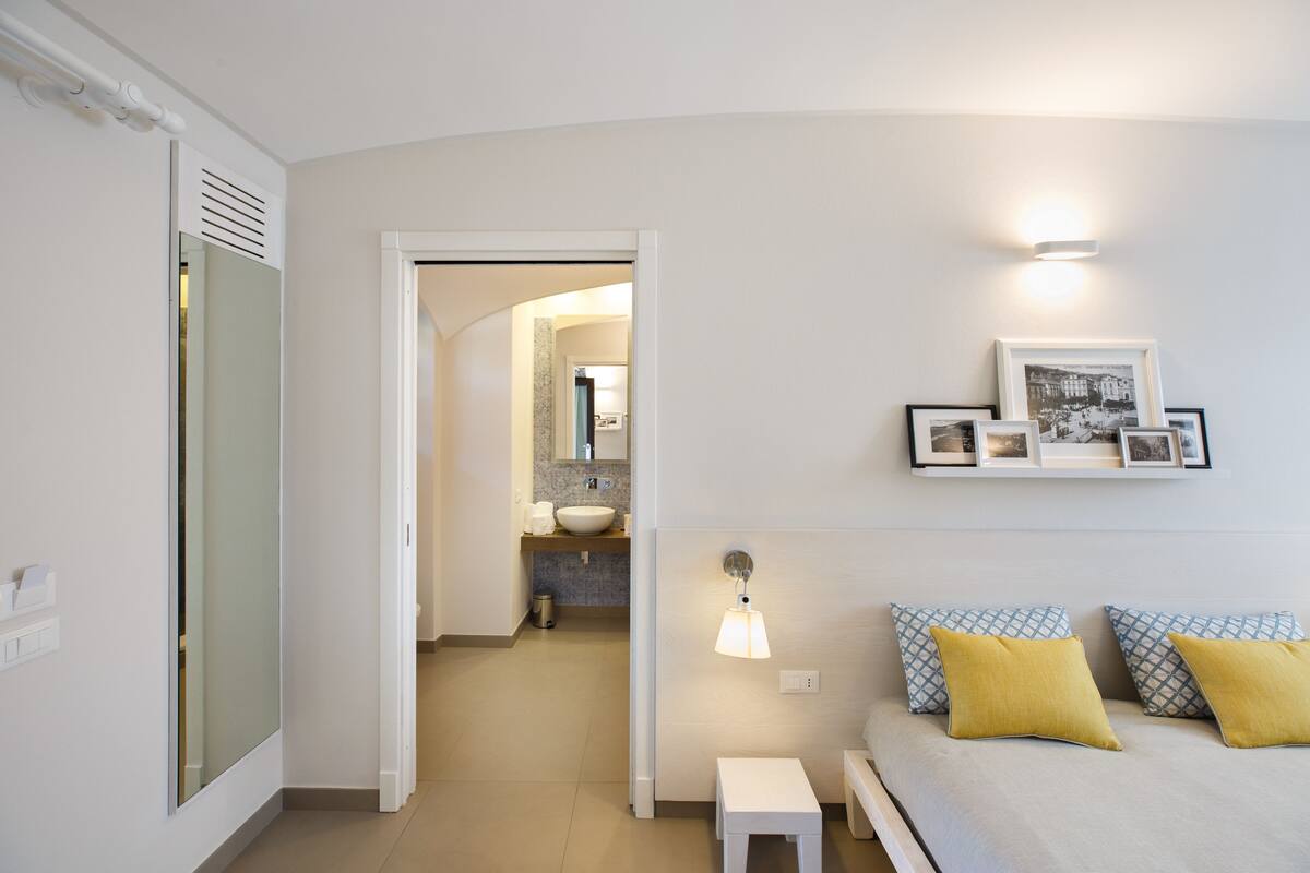 A bedroom with a bed, bedside table, and a wall-mounted shelf with framed photos.
