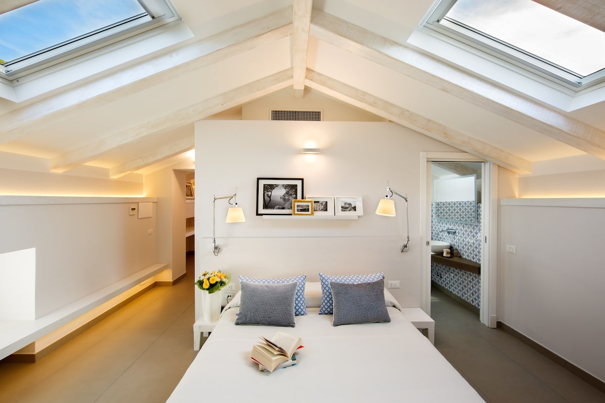 A modern bedroom with skylights, a bed with pillows, a bedside table with books, and a small bedside plant.
