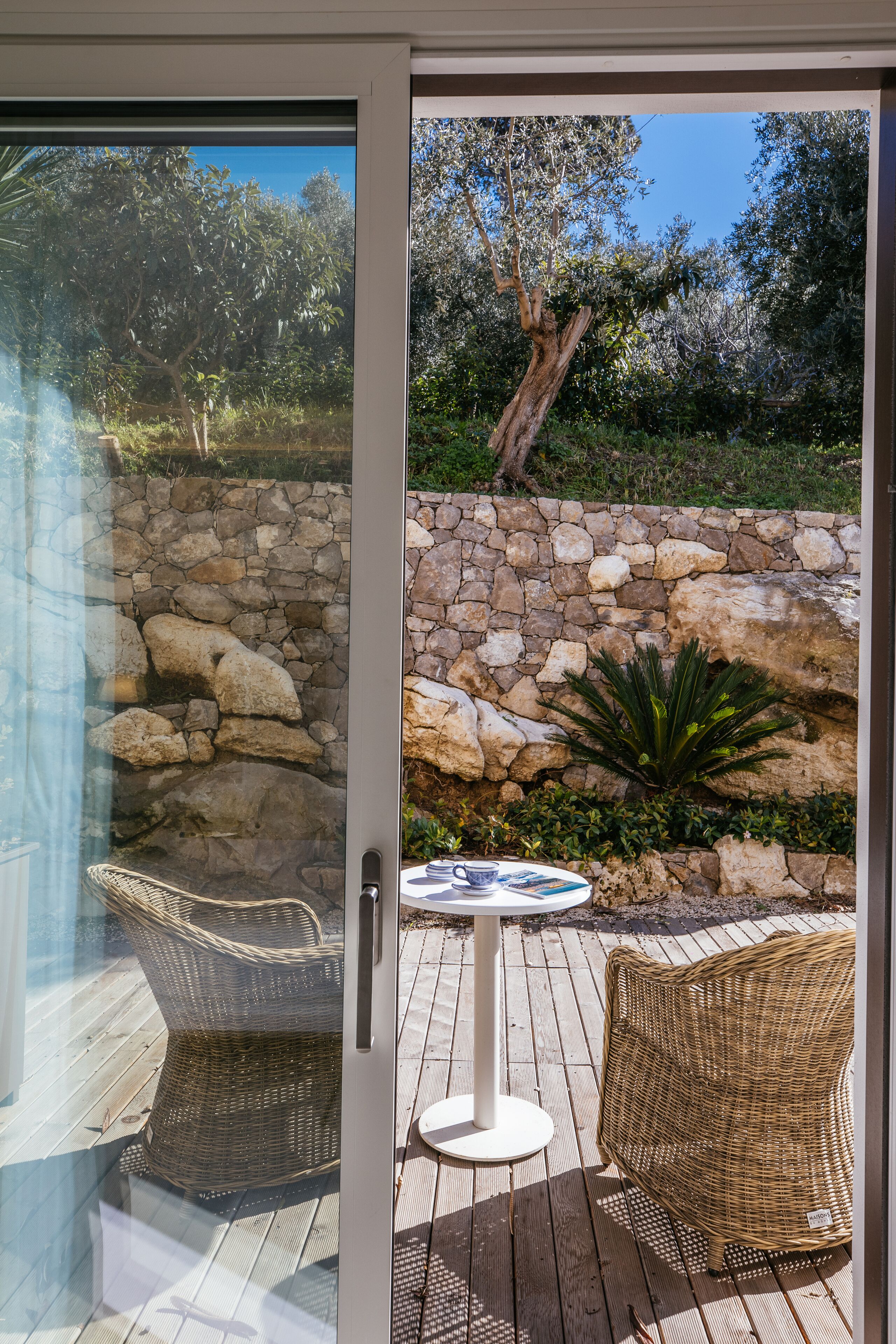 A glass door leading to an outdoor patio with wicker furniture, a stone wall, and a view of the surrounding greenery.