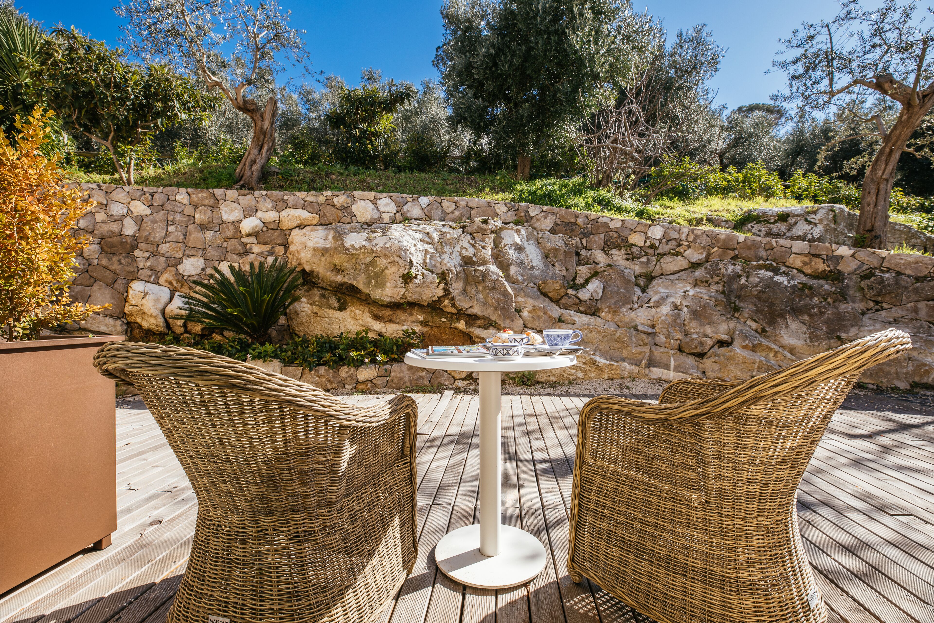 Ein Korbstuhl und ein Tisch auf einer Holzterrasse, im Hintergrund eine Steinmauer und Grünpflanzen.