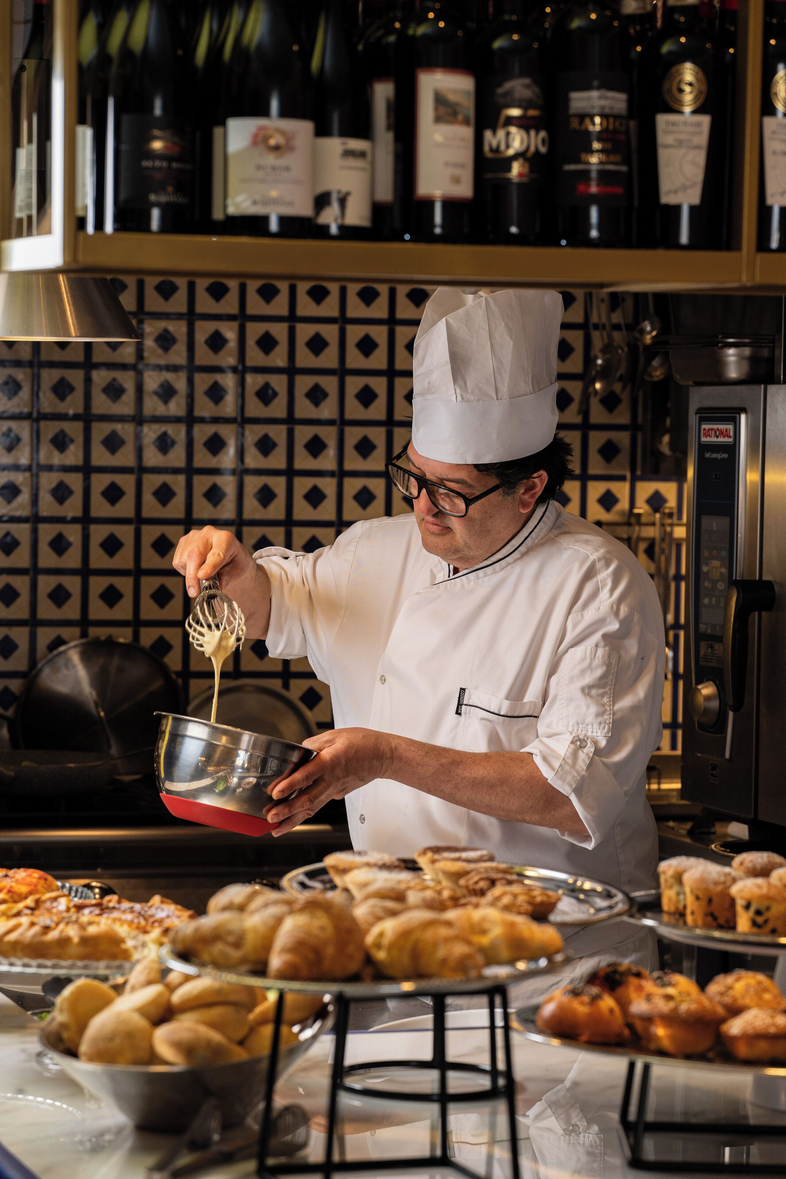 Un chef en una cocina profesional, con diversos productos de pastelería y botellas de vino al fondo.