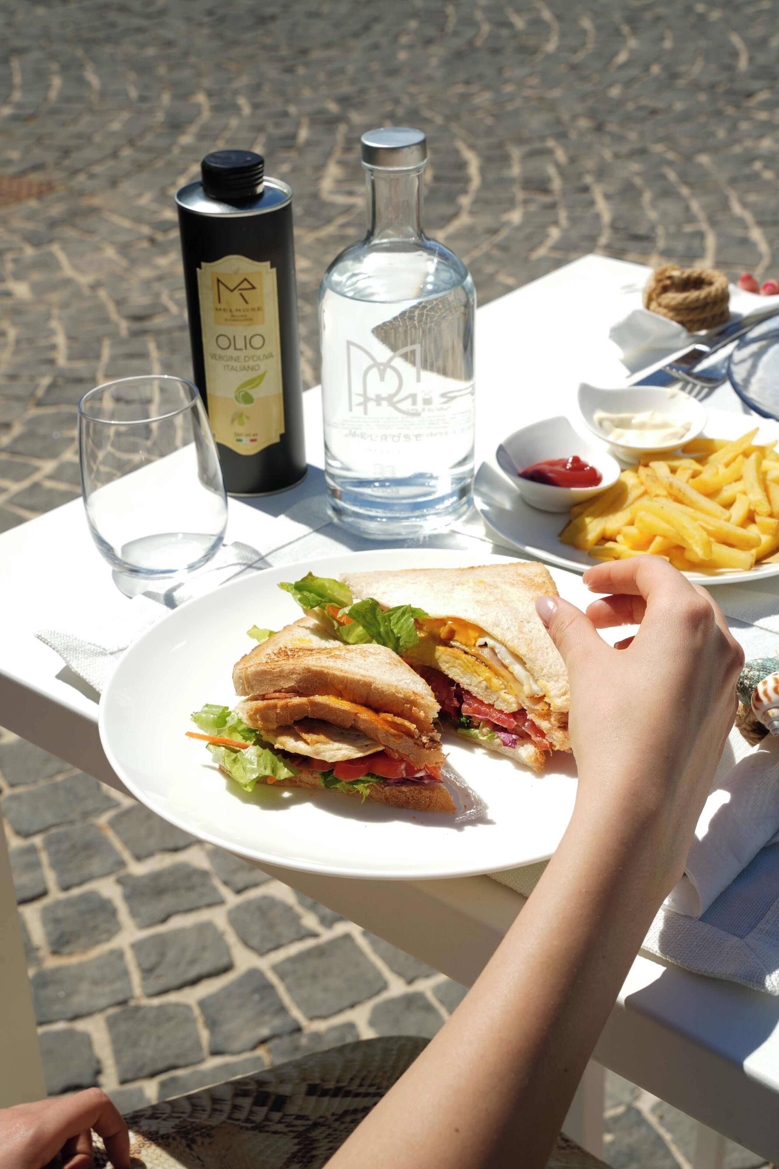Une table avec un sandwich, une bouteille d’eau, une bouteille d’huile d’olive, un verre et une assiette de frites.