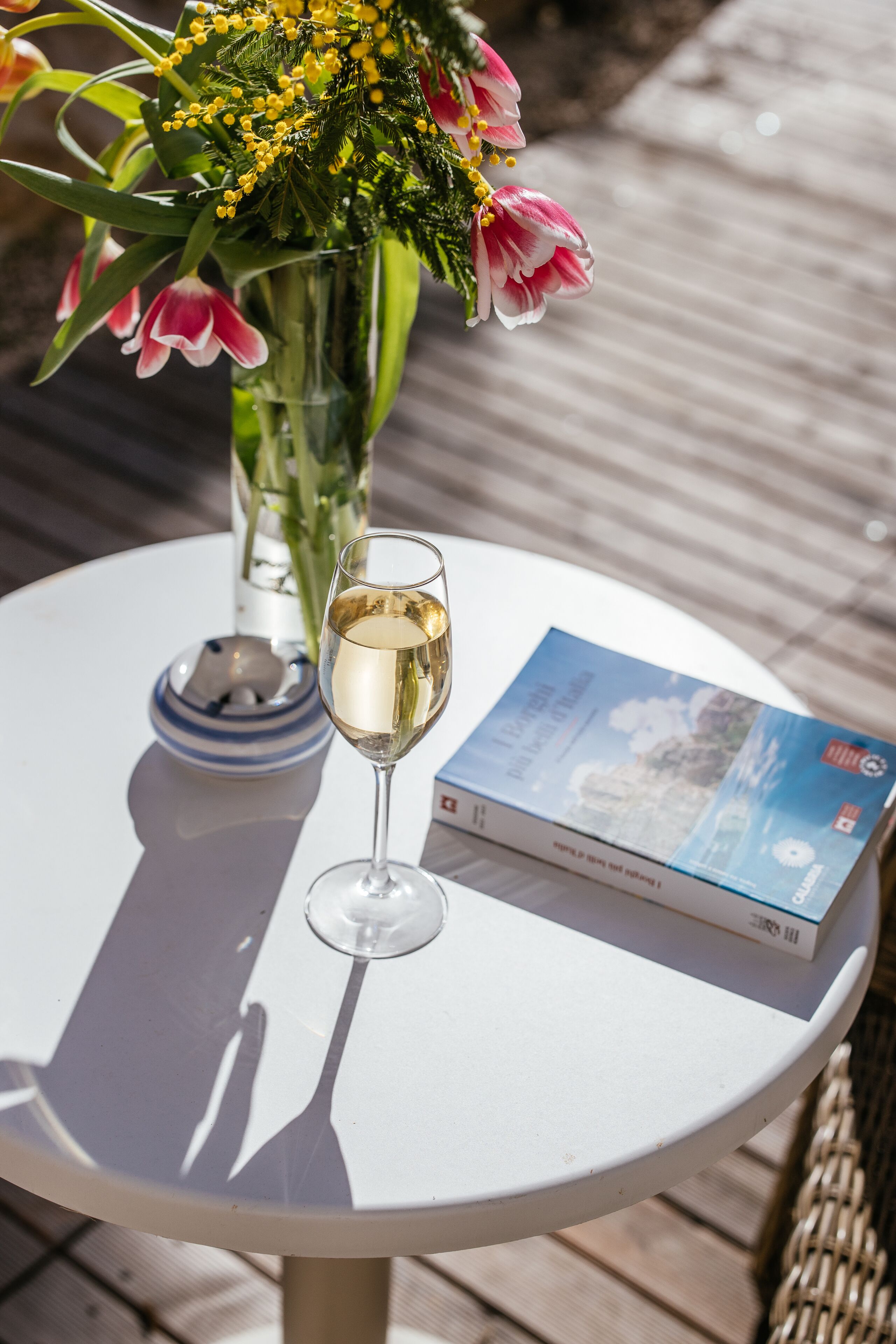 A round white table with a glass of white wine, a book, and a vase of flowers.